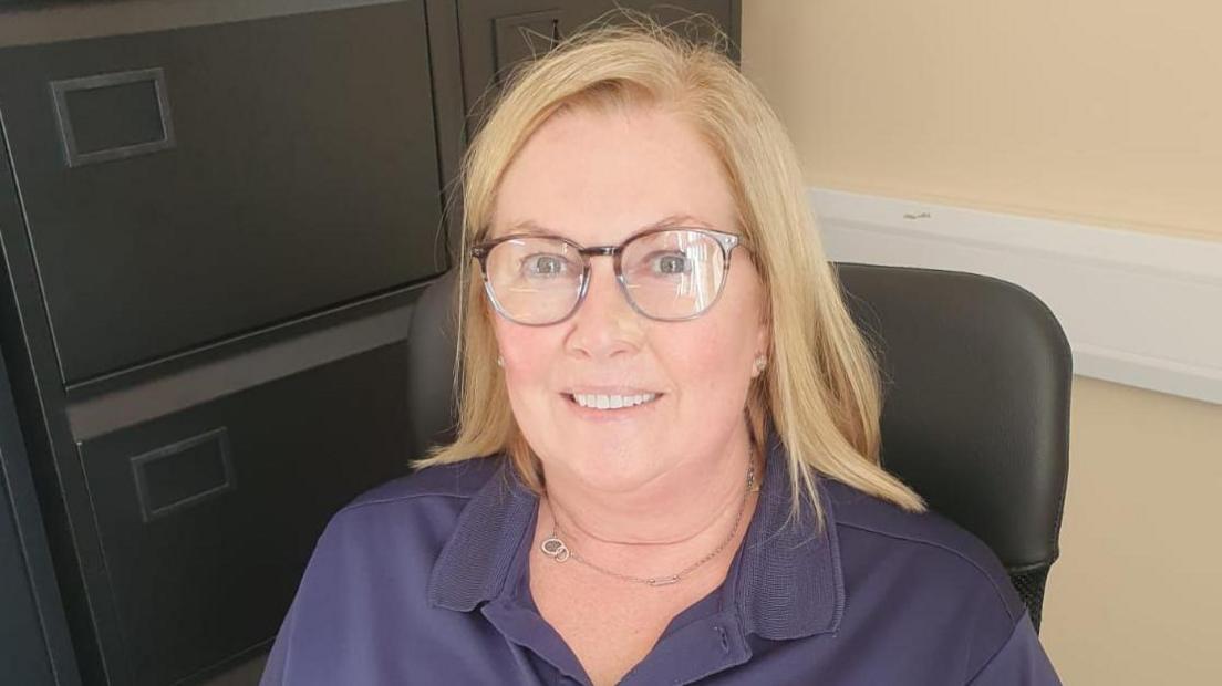 Amanda Logue. She has blonde shoulder length hair, is wearing glasses, a necklace, and a dark blue polo shirt. She is sitting on a chair with black filing cabinets to the left.