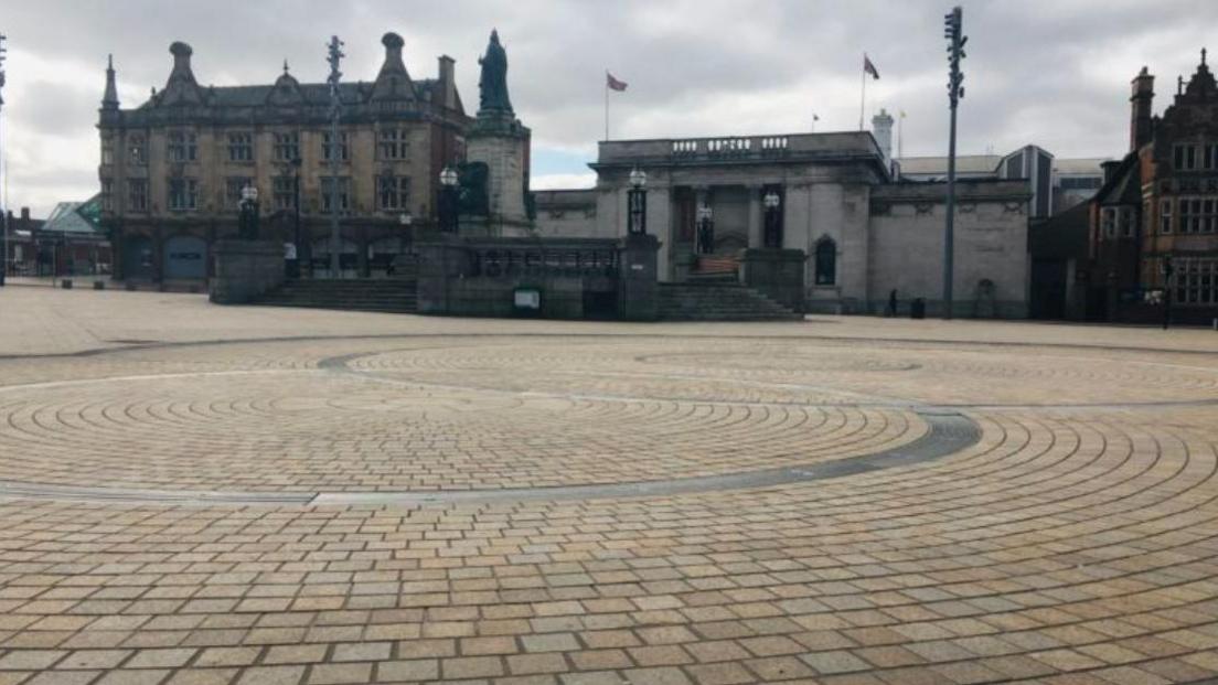 Queen Victoria Square in Hull city centre. Spiralling brick work on the vast space of floor in the foreground with three historical buildings with steps leading up to them in the background.