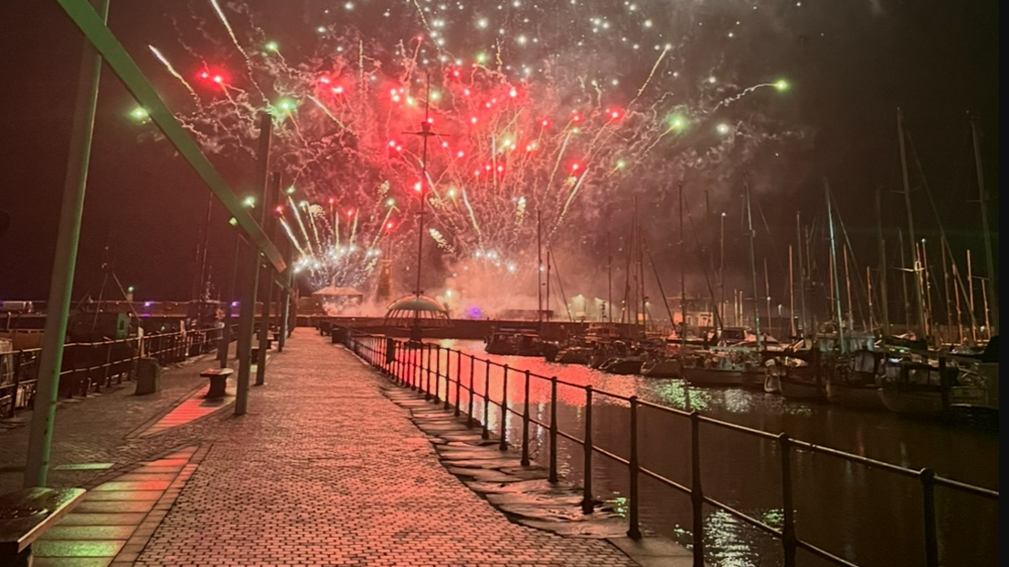 Fireworks over Whitehaven Harbour with boats to one side. The sky is red and gold and white.