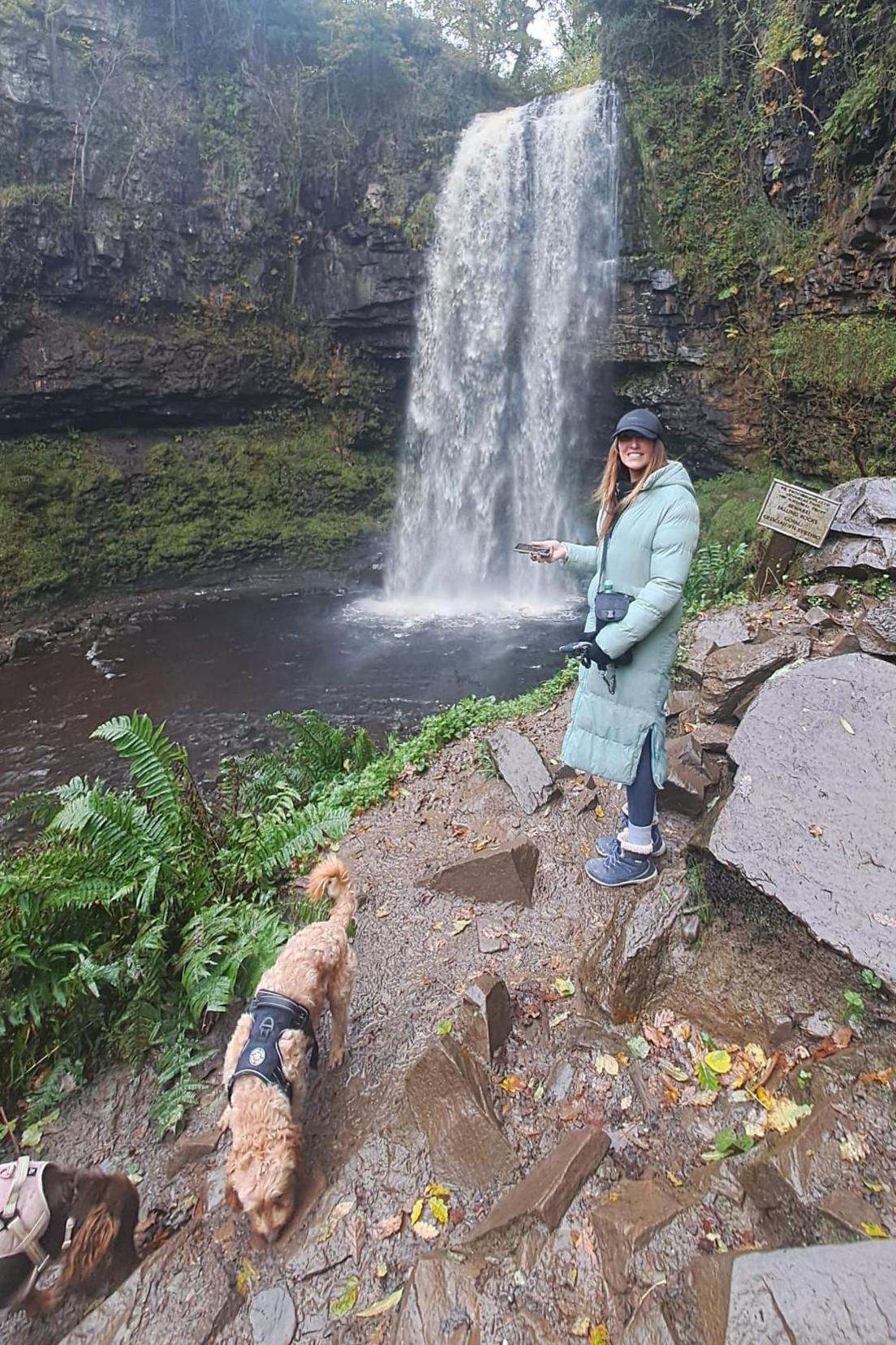 A woman with long bropwn hair wearing a long, light blue blue puffer coat, a black cap and blue walking boots. She is stood in front of a waterfall and surrounded by cliffs. In the forefront of the picture are two small, fluffy brown dogs wearing harnesses.