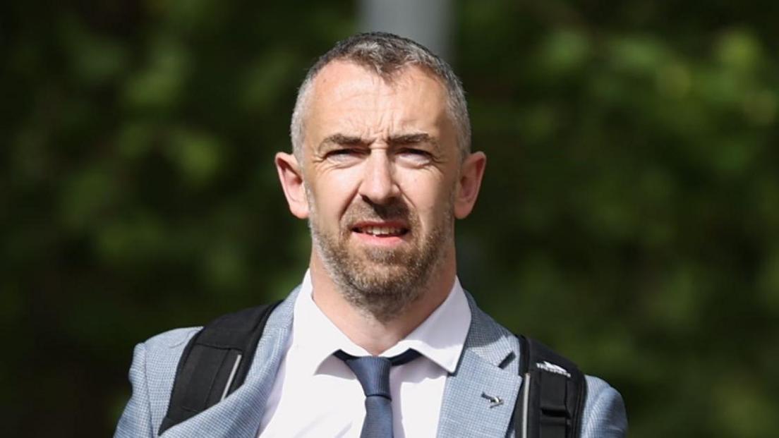 Daithí McKay. He has short salt and pepper hair, wearing a grey blazer, white shirt and navy tie, carrying a backpack.