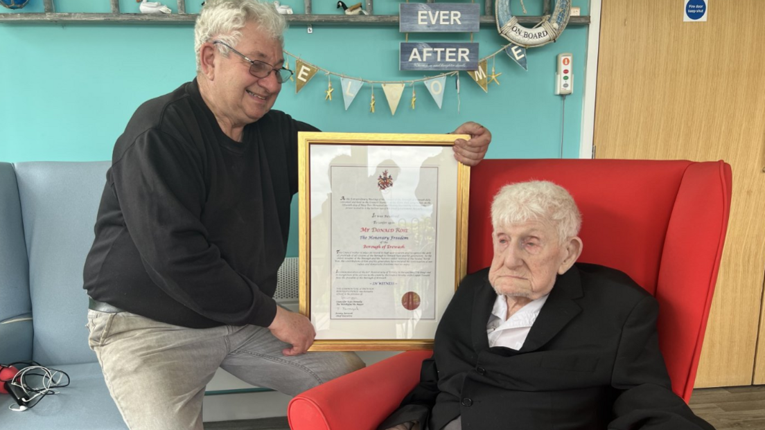 David Rose, who has grey hair and glasses, holds a certificate in a gold frame next to Donald Rose, who is sat in a red armchair.