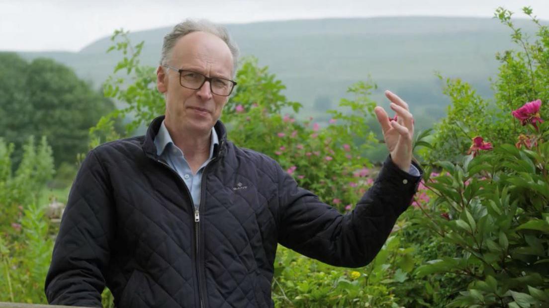 A man in a blue jacket and light blue shirt standing in front of some foliage. He is wearing glasses and his holding his hand aloft.