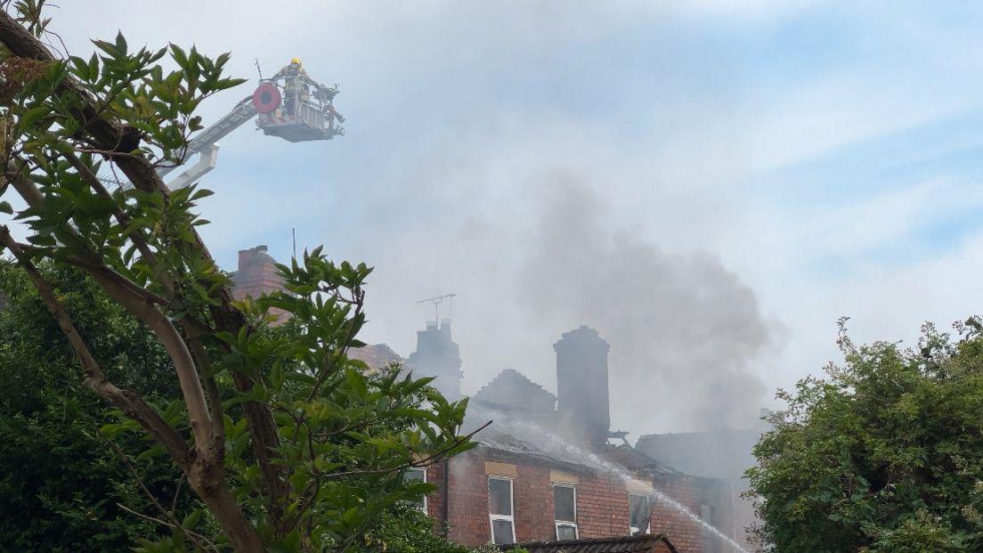 The top of a row of terraced houses. Smoke is billowing from the roof. An aerial ladder with firefighters on can be seen above the houses. Trees with green leaves are in the foreground. The skies are blue.