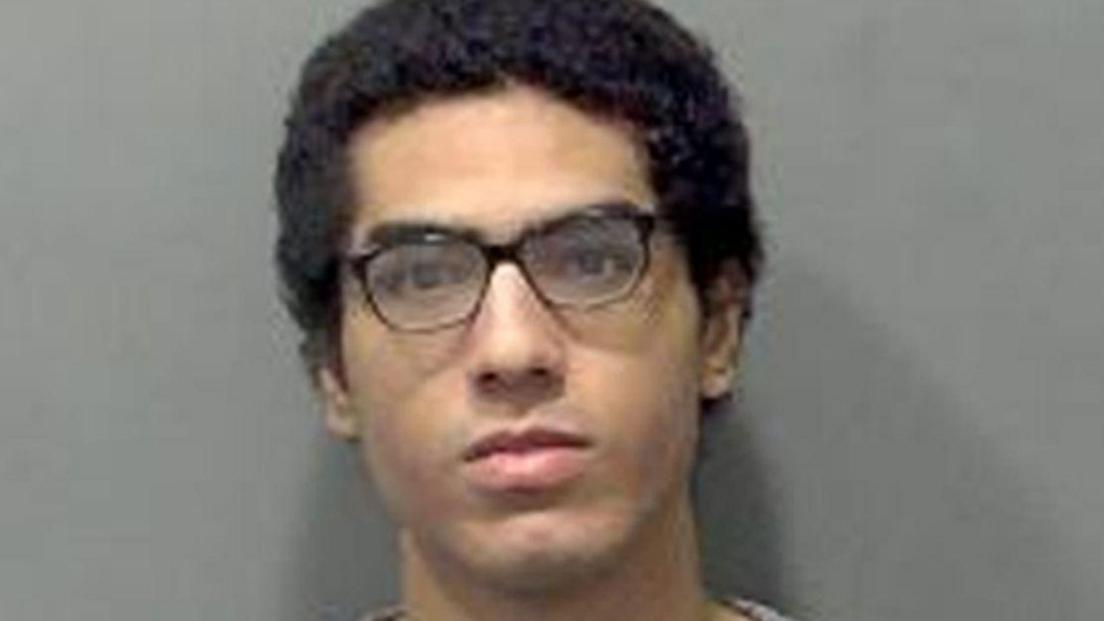 Nicholas Prosper, who has dark hair and dark glasses, staring into the camera for a police custody image