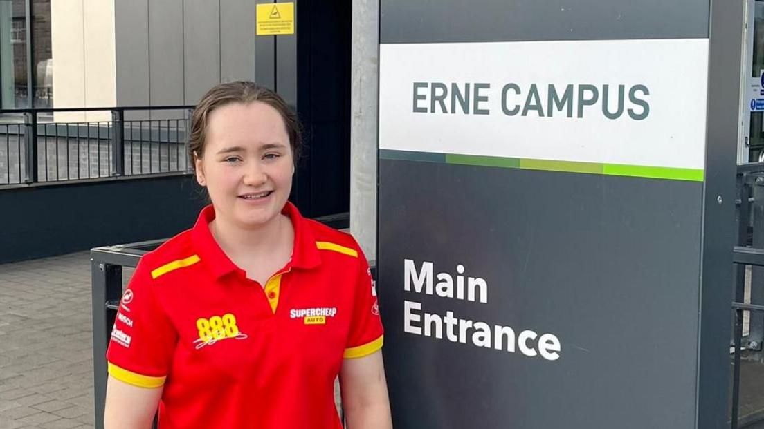 Ella has brown hair in a pony tail. She's standing by a grey Erne Campus entrance sign and she is wearing a red polo shirt with yellow writing on it. 