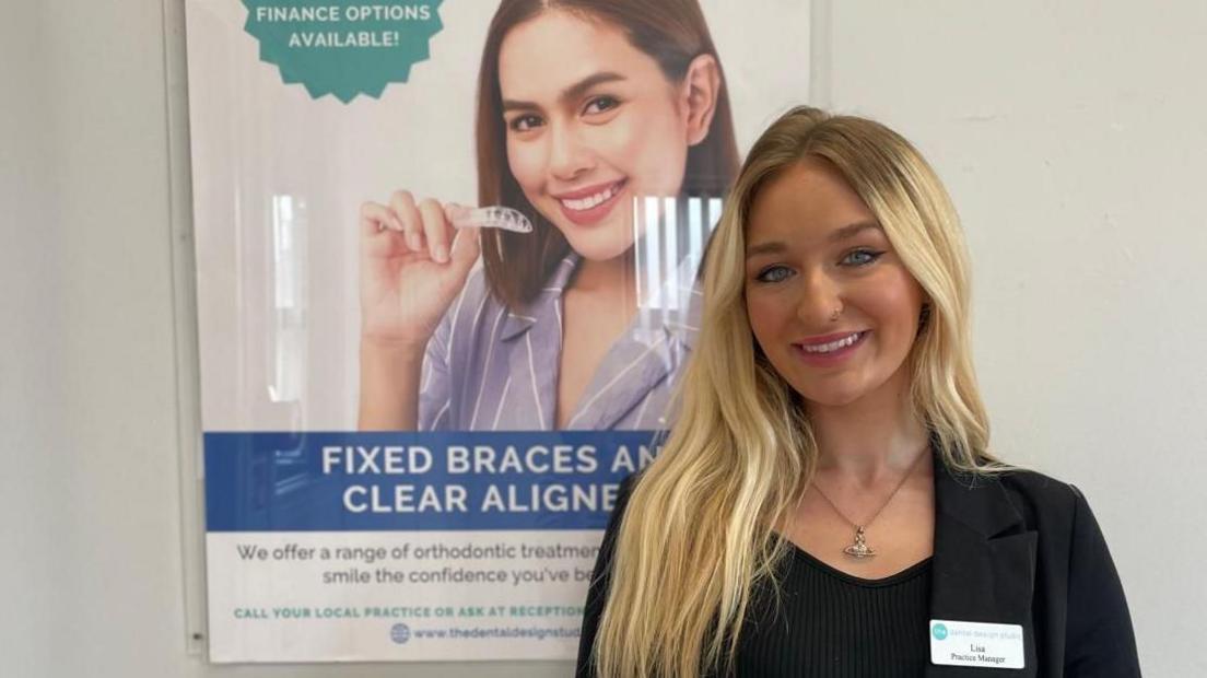 A smiling Lisa Kellie wearing a black top and name badge standing in front of a poster advertising orthodontic treatment