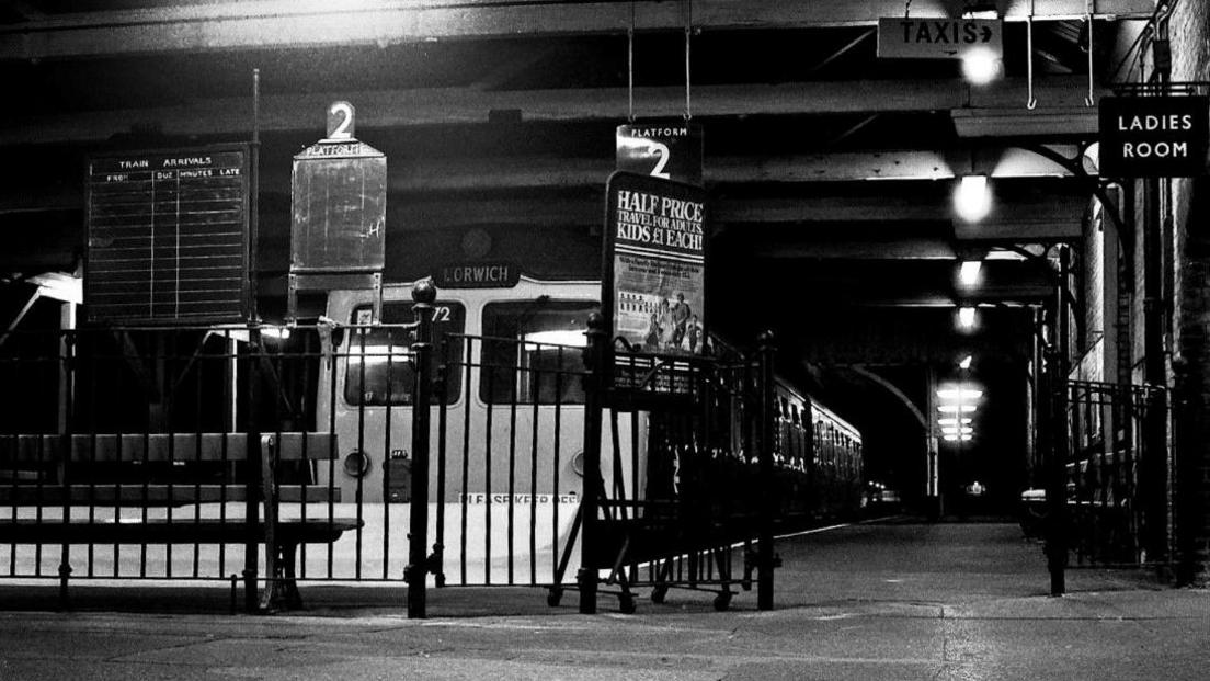 A black and white image of the Lowestoft railway station from decades ago. A train with a sign on the front reading Ipswich rests at a platform.
