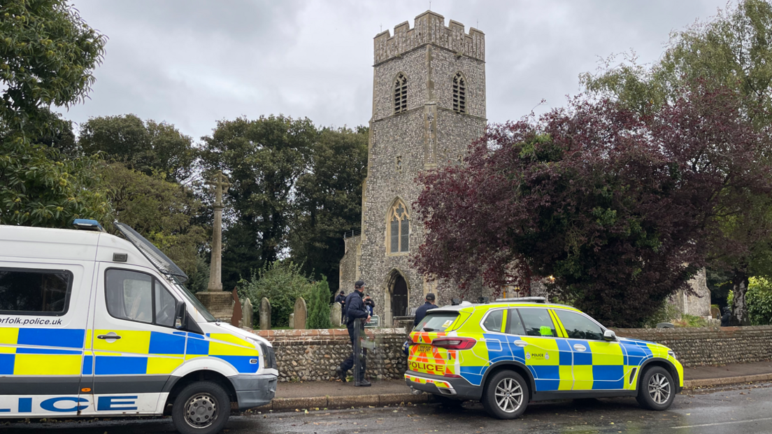 A church with a police van and car parked on the roadside.