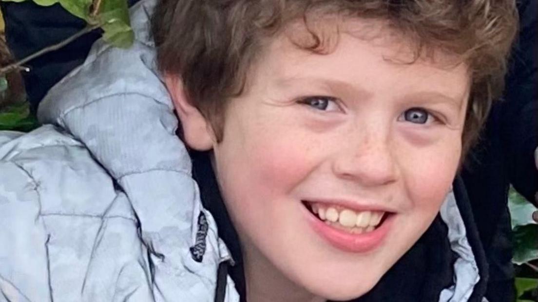 Dylan Cope smiles at the camera, he has curly brown hair and is wearing a grey puffa coat. 