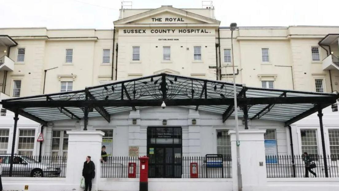 A cream building with The Royal Sussex County Hospital written on it in black. There is a black metal portico at the front of the building, and a low white wall