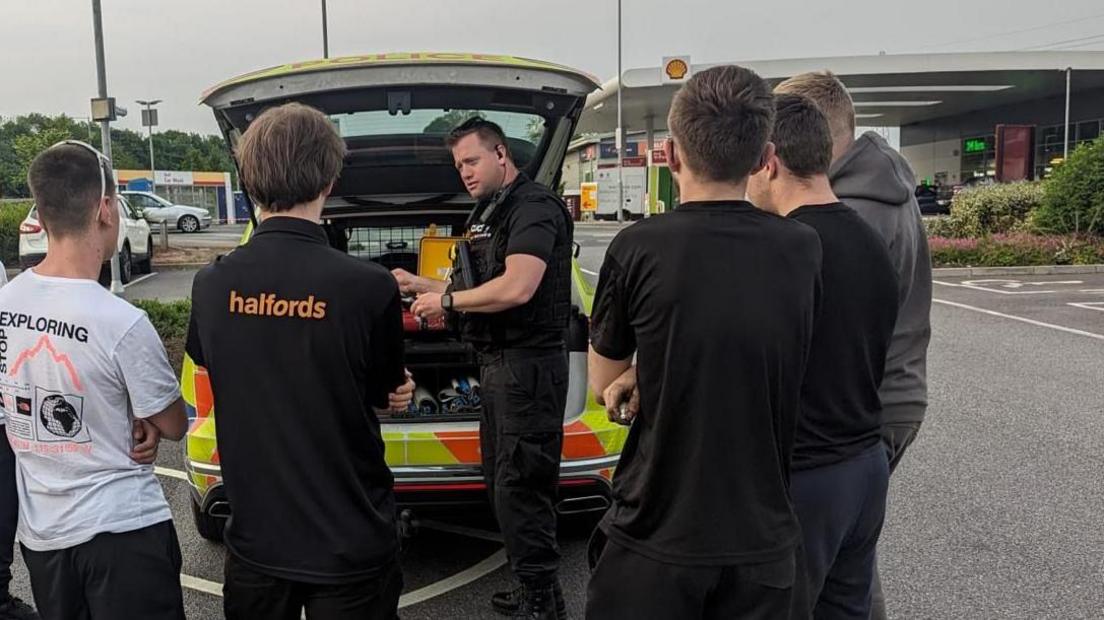 A police officer dressed in black standing in front of his police car, which is parked in a petrol garage with the boot open. He is talking to a group of young men who have their backs to the camera.