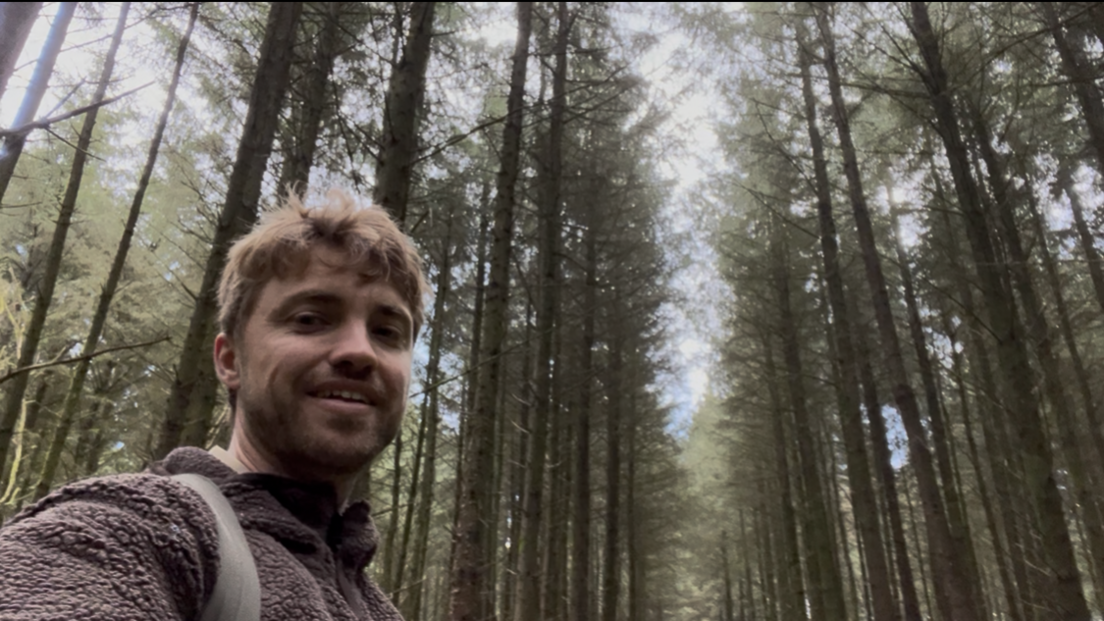 Max Weston in a selfie, smiling down at the camera with tall trees in the background and sky behind them. He is wearing a brown fluffy jumper and has rucksack straps over his shoulders.