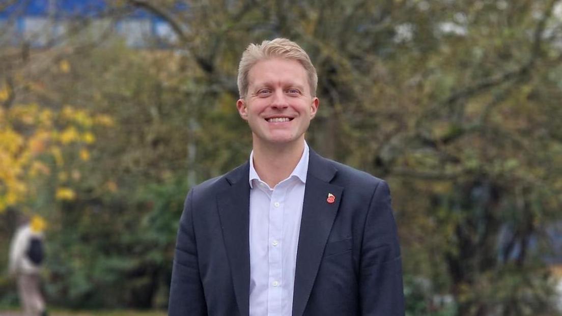 Dr Ben Spencer smiles at the camera, he is wearing a dark blue suit jacket with a white shirt underneath with the top button undone. In the background out of focus trees are seen