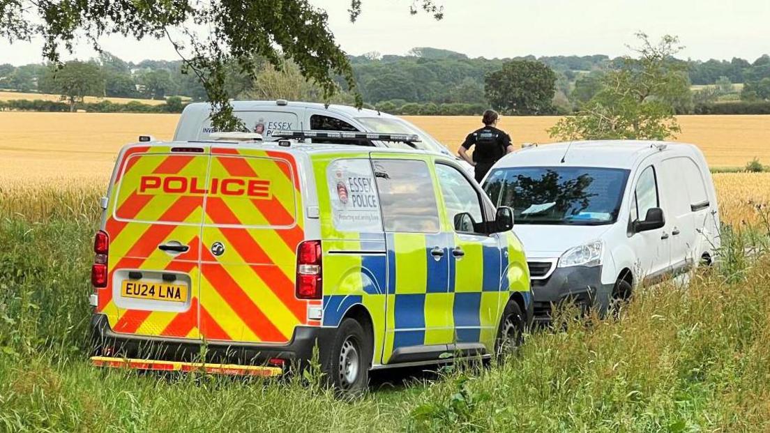 Three police vans parked on a field, with an officer looking away into the field.