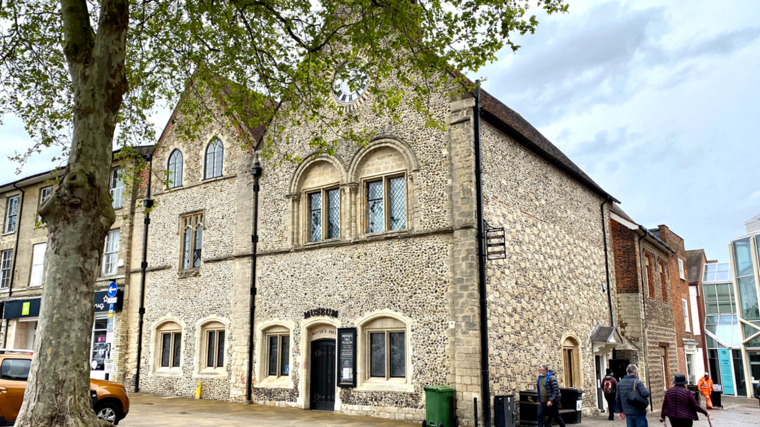 The front of a museum in Suffolk shows an old stone building with ornate arched windows and a clock on the front. People are walking past it on the street