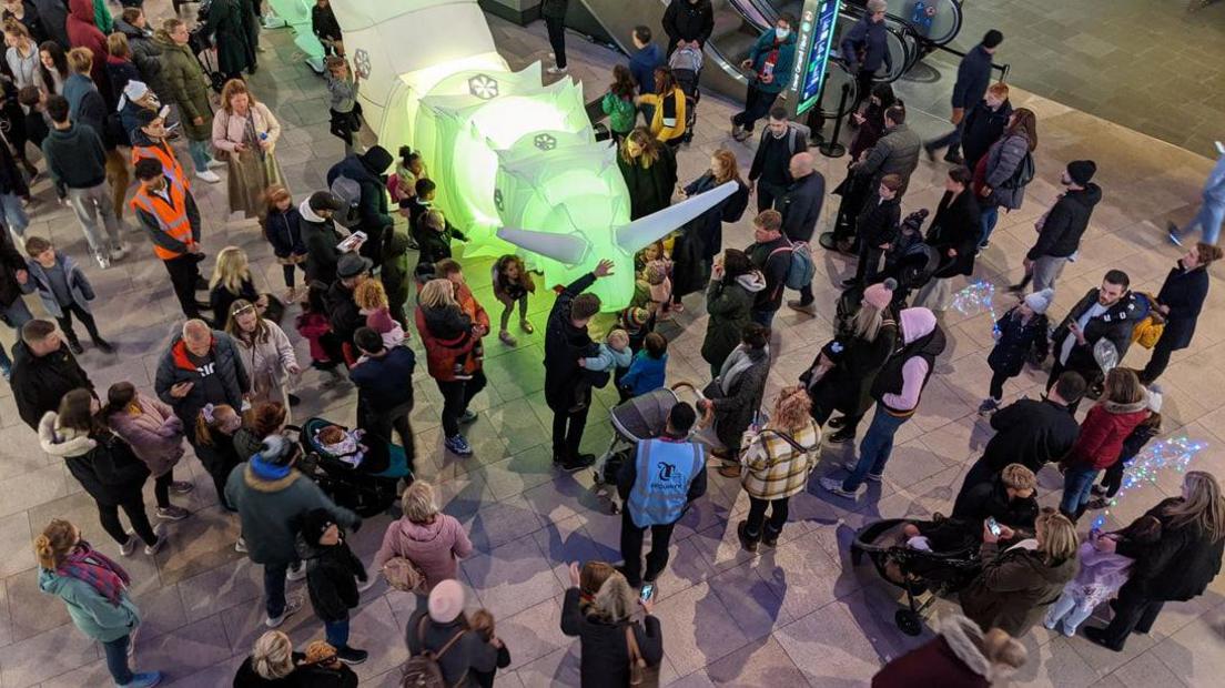 Overhead view of a large crowd of people around the illuminated snail in a museum hall as a man holding a child reaches out to touch the inflatable robot.