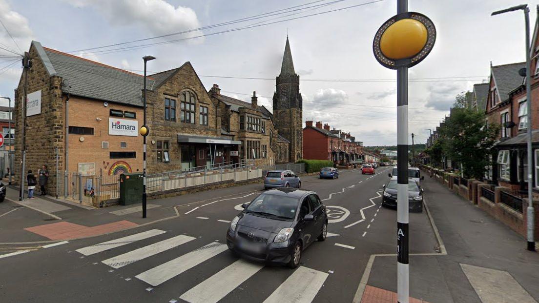 A street view image of the road shows a row of buildings on either side, ranging from a church to houses. Several cars are parked on the side of the road. A black Toyota car is in the process of driving across a zebra crossing.