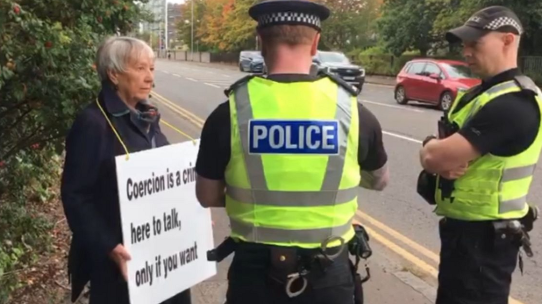 Rose Docherty stands on a pavement holding a protest sign that reads, "Coercion is a crime, here to talk, only if you want." She faces two uniformed police officers wearing high-visibility jackets with "POLICE" written on the back. One officer has his arms crossed while the other appears to be speaking to Rose Docherty. A red car and greenery are visible in the background.