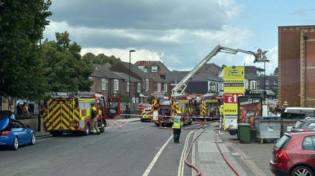 Three fire engines stopped on a cordoned-off road. There are hoses laid out on the pavement.