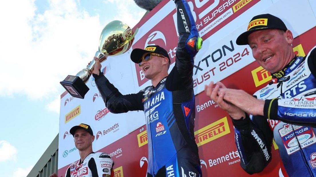 Ryan Vickers is lifting his arms and hold a large gold trophy. He is wearing blue and black-coloured racing OMG Racing Yamaha gear and a black and yellow cap. There are men either side of him also wearing black and yellow caps. They are standing in front of a large white, red and yellow advertising hoarding.