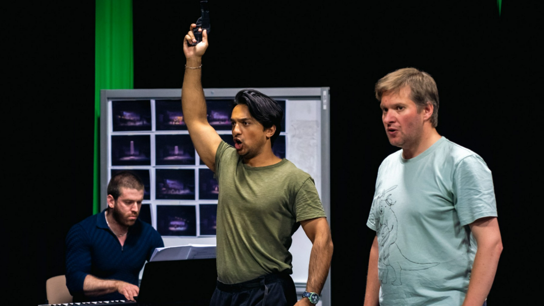 Three performers on stage. Two of them are standing in the foreground and one has his arm raised as he points a pistol in the air. A third person is sat down and playing a keyboard