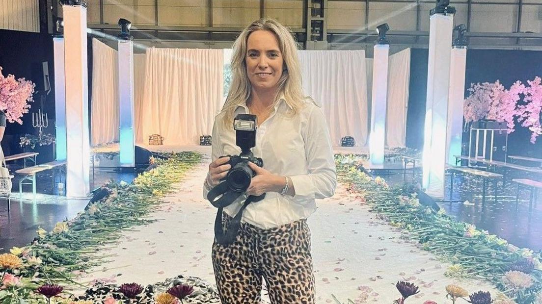 A lady standing in front of a catwalk that has flowers laid along the edge. She is wearing leopard print trousers with a white shirt and is holding a professional camera. The lady, who has shoulder length blonde hair is smiling. 