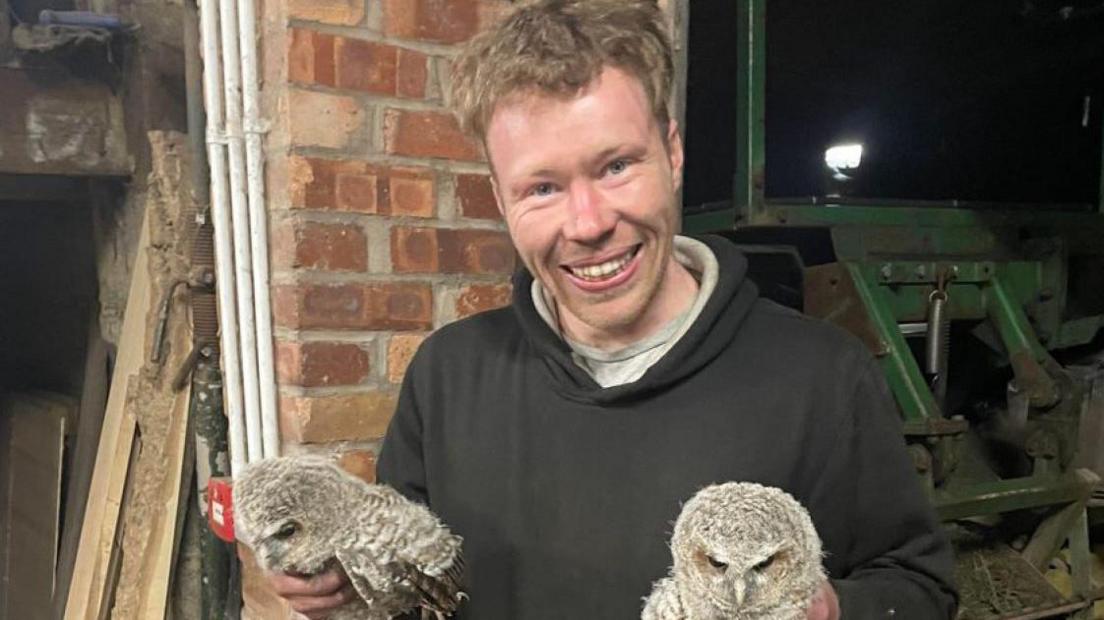 Carruthers, who has short fair hair and is wearing a black hoody, holds two young grey owls. He is smiling with agricultural equipment behind him