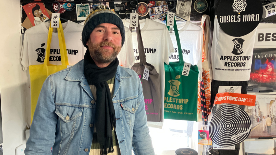 A man in a woollen hat, blue denim jacket and black scarf looks at the camera. Behind him are T-shirts and bags with the Applestump Records logo on them.