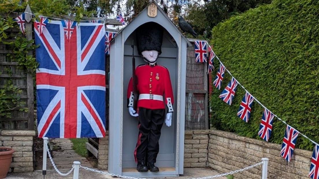 A scarecrow King's Guard soldier standing to attention in his guard hut surrounded by the union flag and bunting