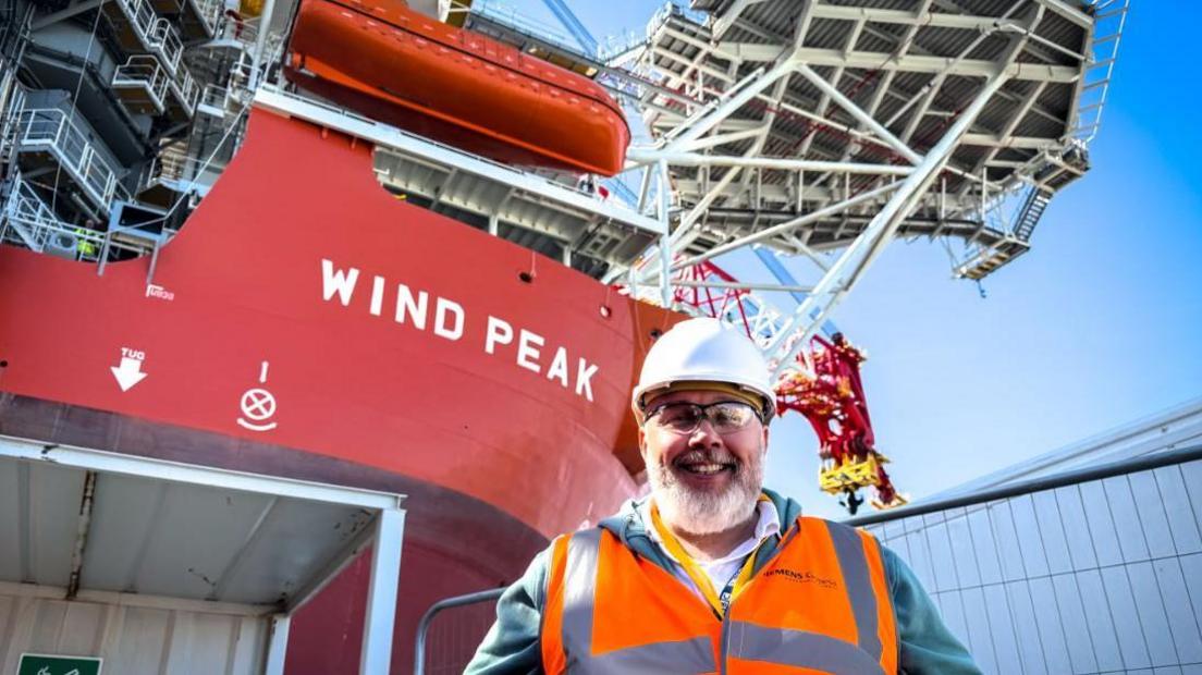 A smiling man stands on a ship, wearing glasses, a hard helmet and a hi-vis. Behind him rising a red superstructure, with the words WIND PEAK written on it in white letters.
