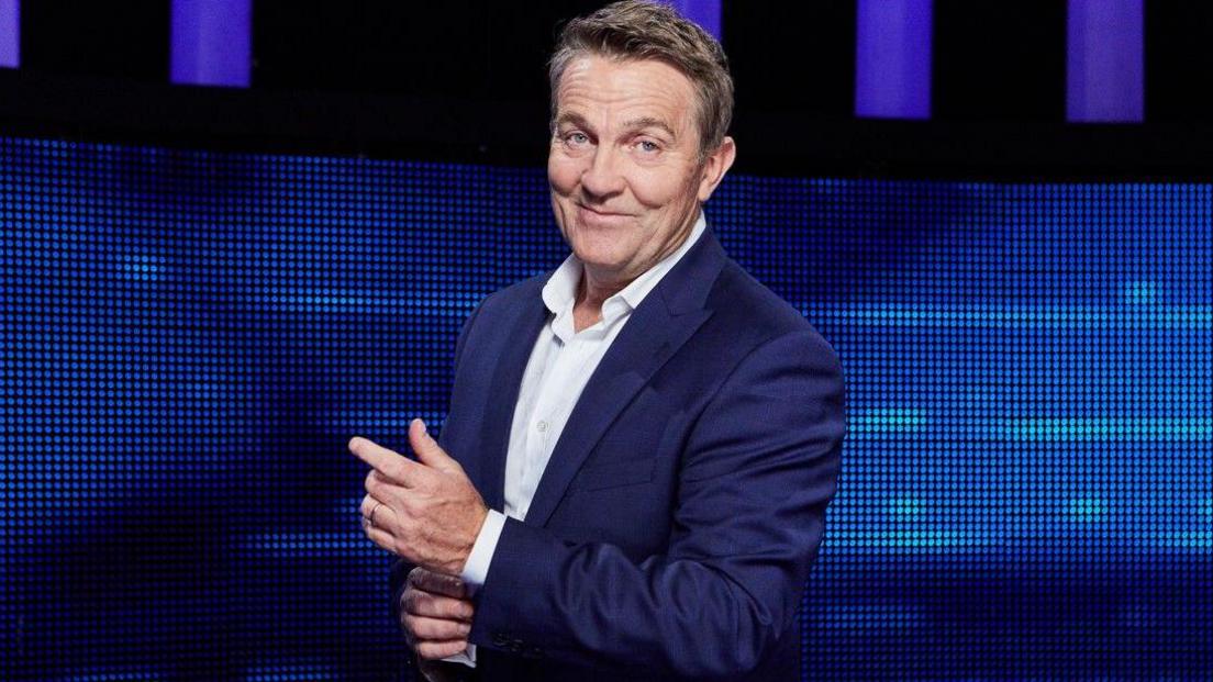 Bradley Walsh in dark blue suit with white open-necked shirt.