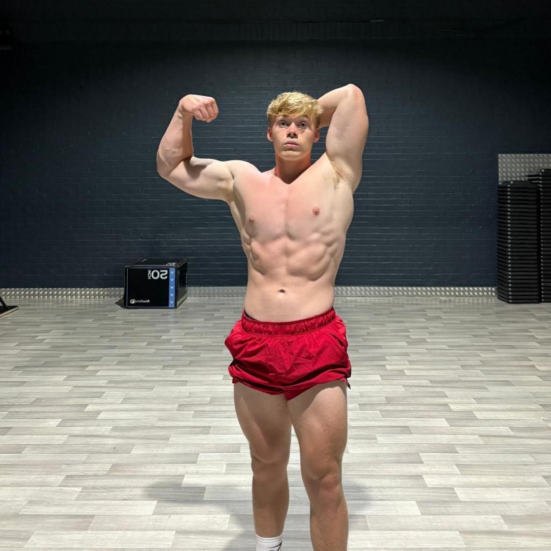 George Holland a teenager poses for the camera in a body-building stance, wearing red shorts and flexing.