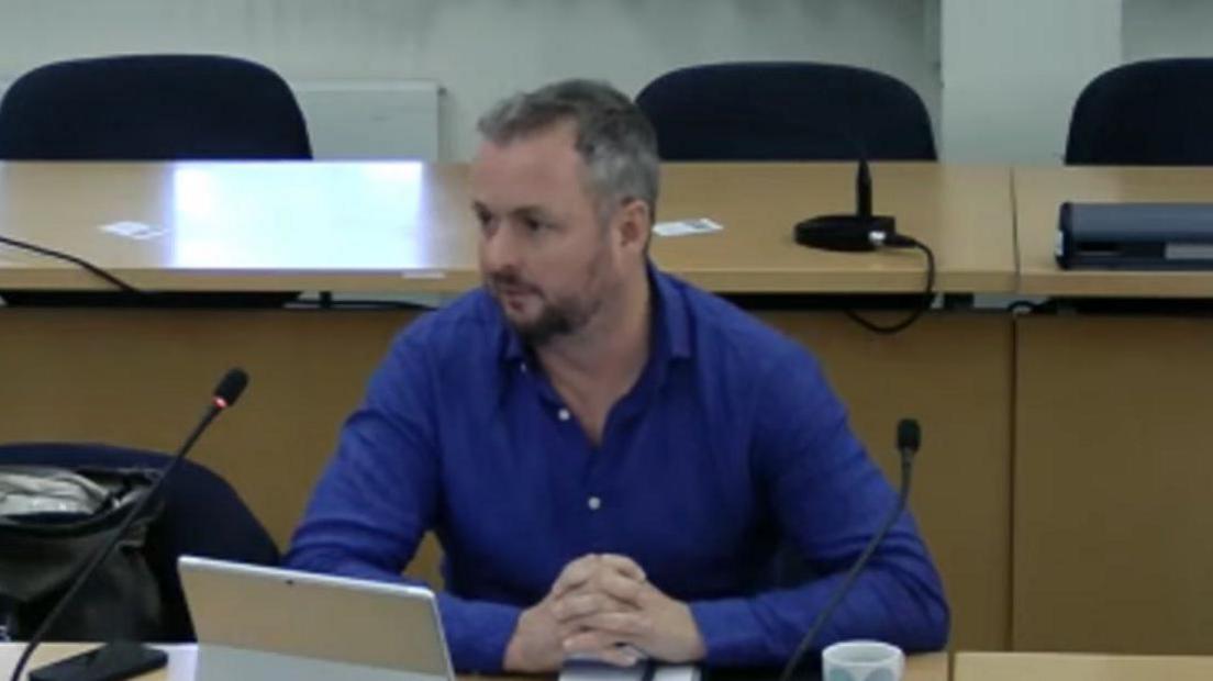 A man is sat in a council chamber with a microphone pointing towards his mouth and a tablet computer on the desk in front of him. He is wearing a blue shirt.
