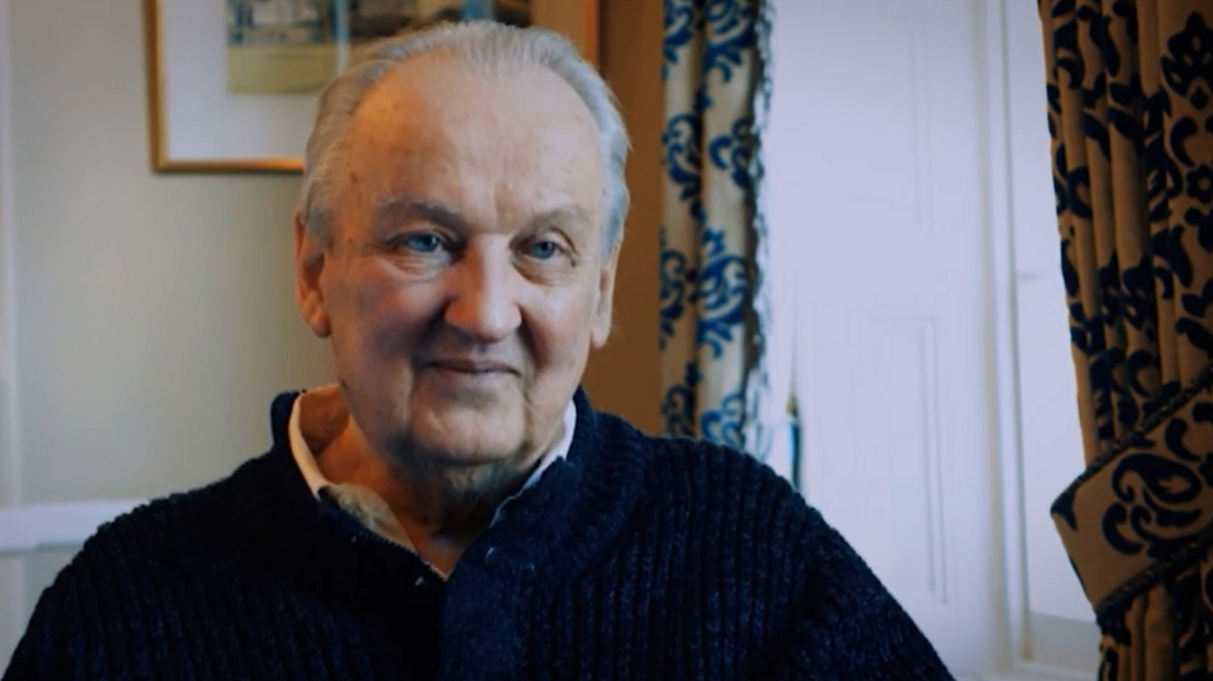 Douglas Gowan wearing a dark knitted jumper sits indoors beside a bright window. Patterned curtains frame the window, while a framed picture hangs on the wall behind. The photograph is warmly lit and focuses closely on his face and upper body.