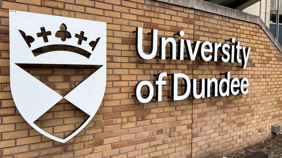 A white sign on a brick wall with a crest and the words University of Dundee