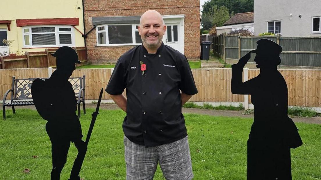 Dave Hubbard is standing beside the two statues in on the grass. He is wearing a chef's tunic and grey chequed trousers