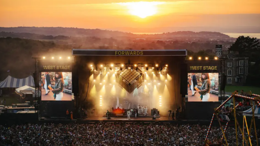 A promotional image of a large music festival stage, and two smaller TV screens, projecting images of bands, in front of a huge audience on the Downs. It is dusk and the sun can be seen setting in the distance behind clouds