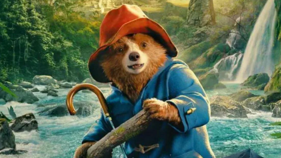 Paddington bear wearing a blue anorak and red hat is holding a tree log which he is presumably using to row in a boat down a fast flowing rainforest river with waterfall in the background. The image is taken from the film Paddington in Peru 