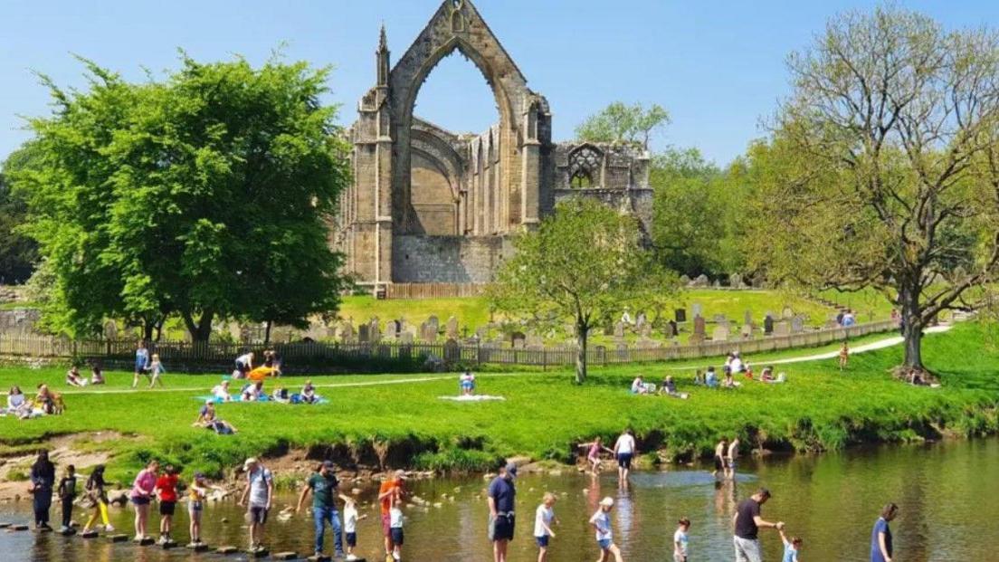 A sunny day at The Bolton Abbey estate in the Yorkshire Dales. Visitors are lying on the grass in the sun while others are waiting their turn to get across the stepping stones.