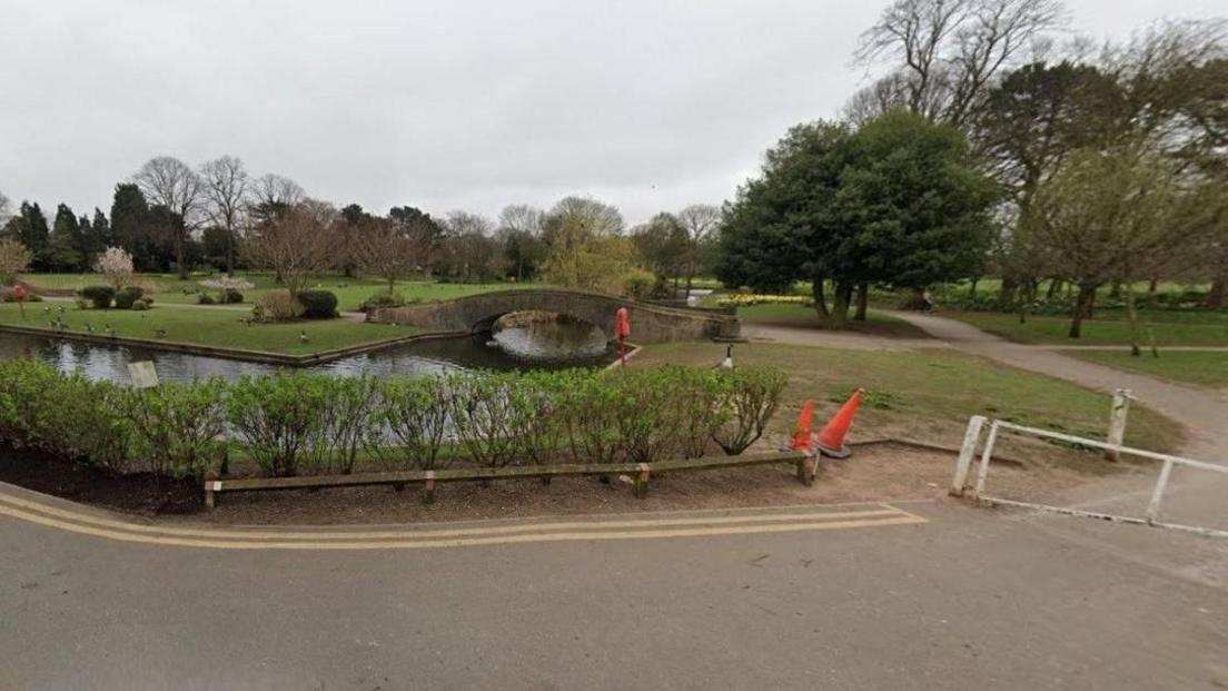 A park on a cloudy day. There is a river with a low footbridge surrounded by grassy areas. There is a tree-lined path on the right.