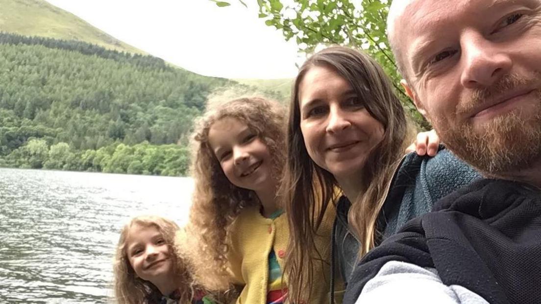 The family are lined up, youngest to oldest - Ivy, sister Lydia, 12, mum Cherry and dad Stuart smiling at the camera beside a lake. There are lots of trees across the water. 