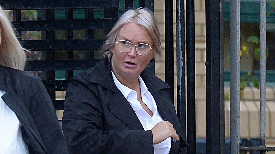 A woman exiting a turnstile. She is wearing a black coat over a white blouse and has cropped blonde hair and glasses.