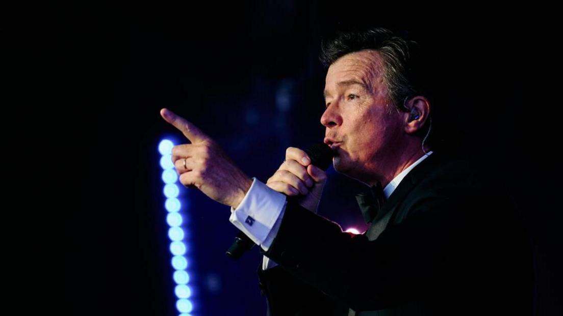 A man with dark hair and a dark suit in the dark, holding a microphone