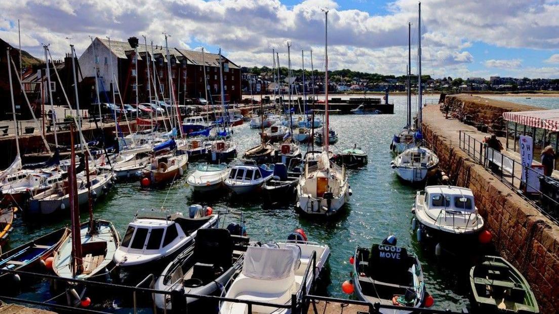 A colourful harbour filled with sailboats and small motorboats moored along stone quays. Bright blue skies and large white clouds overhead add contrast to the bustling waterfront and the buildings lining the harbour.