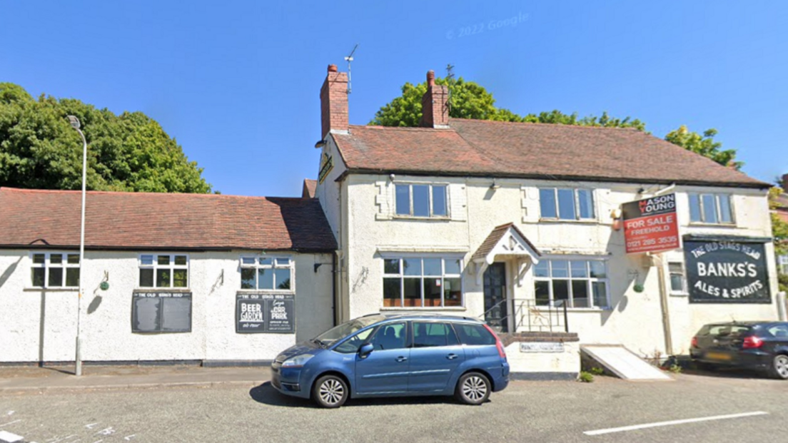 The pub is white and has an adjoining building. There is a for sale sign up with two cars parked outside the front.