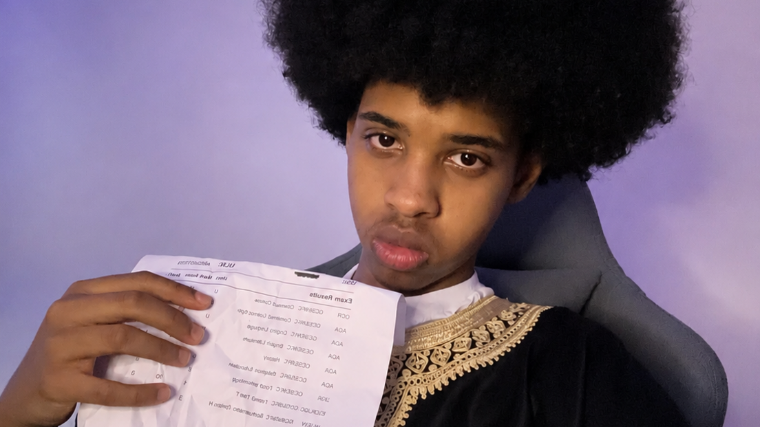 Adbi, with acheronian afro-style hairsbreadth holds a crumpled printed papers and wears a acheronian tunic with golden embroidered trimgainst a partition lit by bluish LED lights.