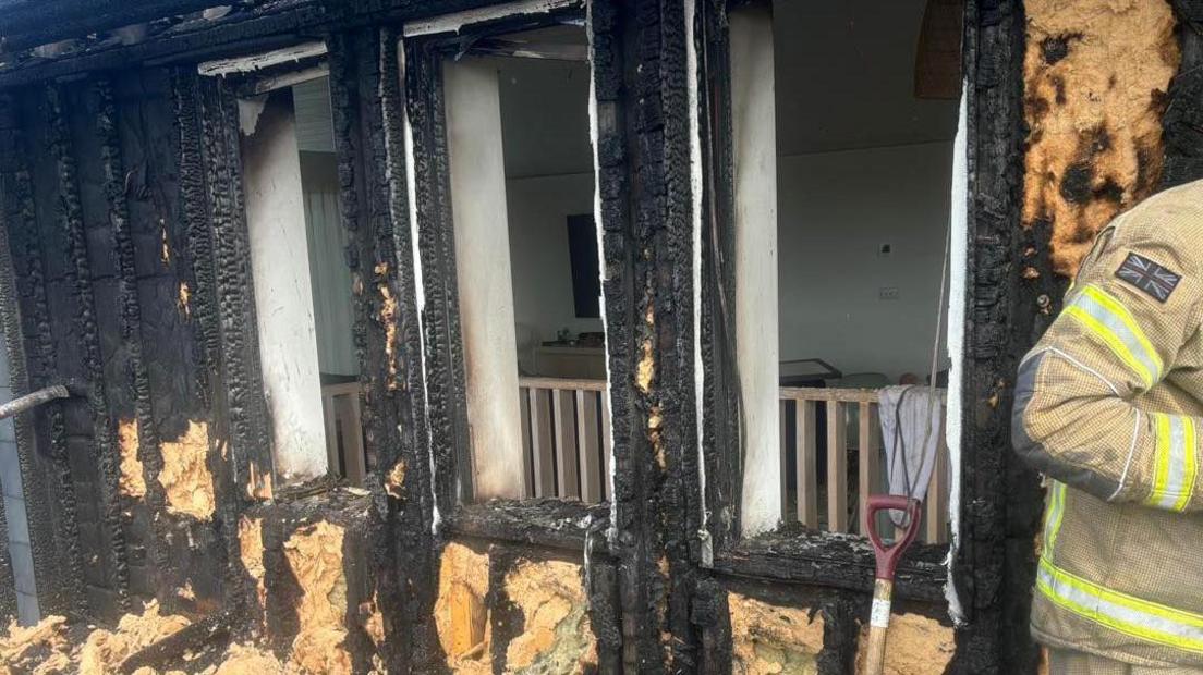 Charred exterior walls which have been turned black following a fire in Cornwall. Part of a firefighter in uniform can be seen on the right-hand side of the photo and the top half of a shovel is propped against one of the walls.