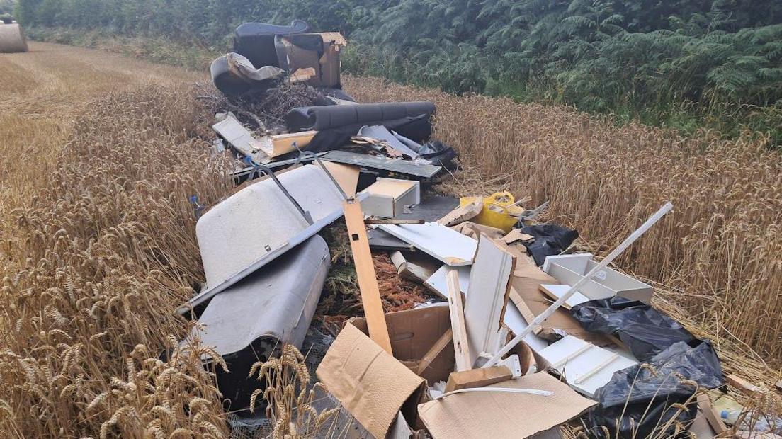 A pile of rubbish including a bath and pieces of wood on the ground in a field. There are trees in the background. 