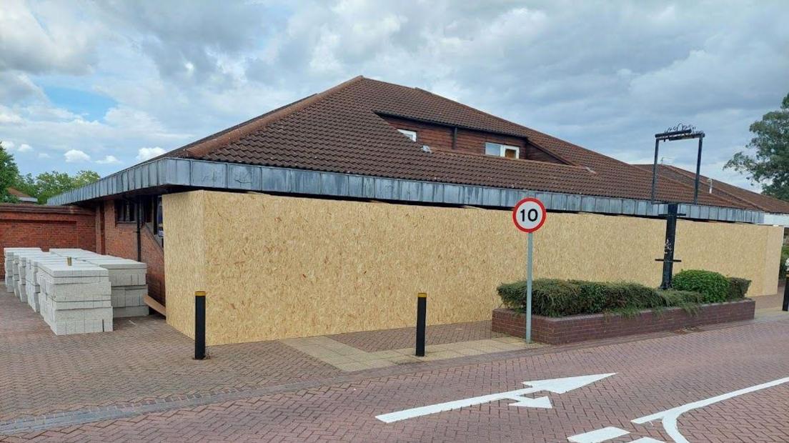 A red-brick building with light brown boarding across its entire front. There are pallets of what look like concrete blocks stacked to the left of it.